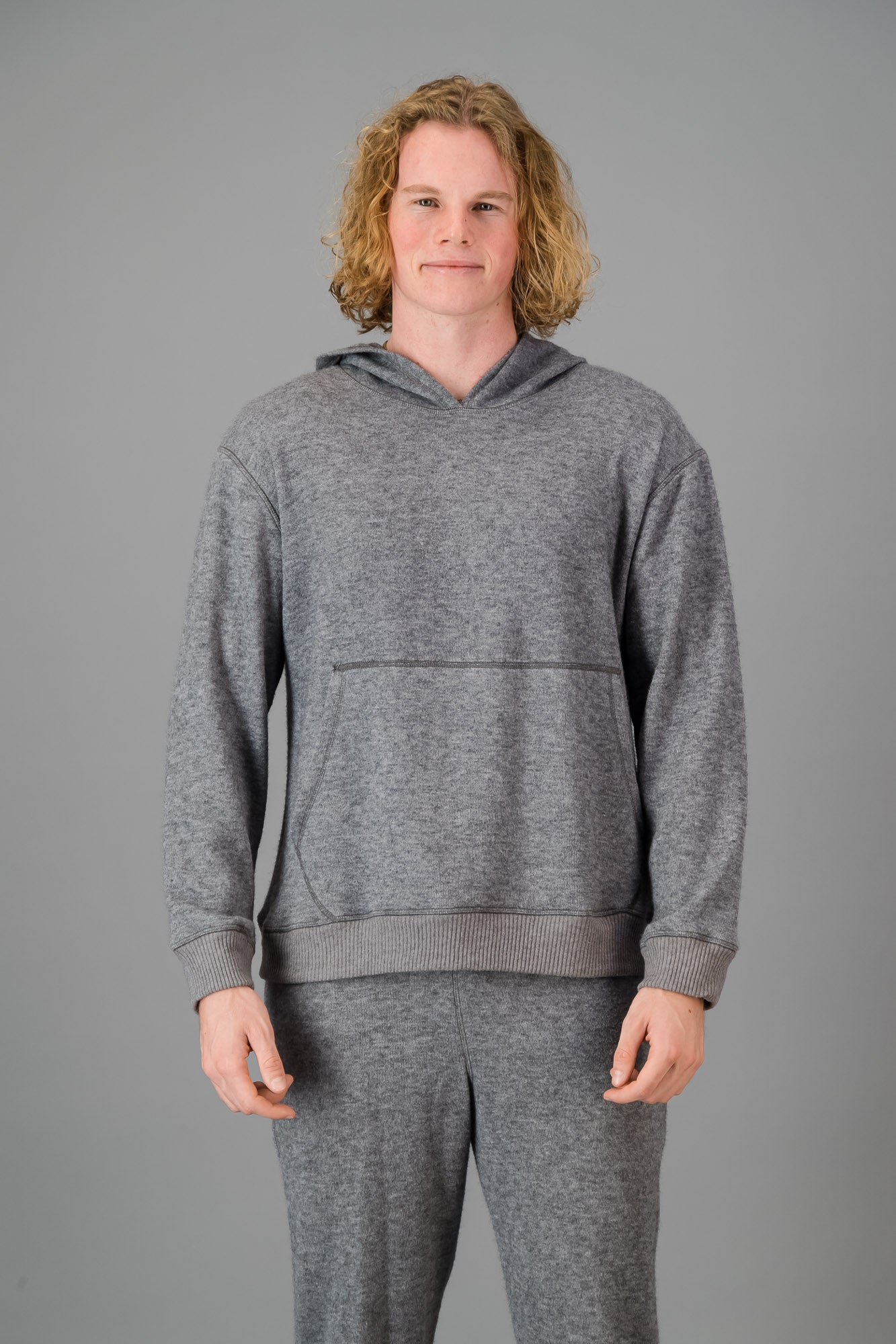 Person with curly blonde hair wearing a comfortable Cashmere-Feel Hoodie (Grey) and matching relaxed fit grey joggers.