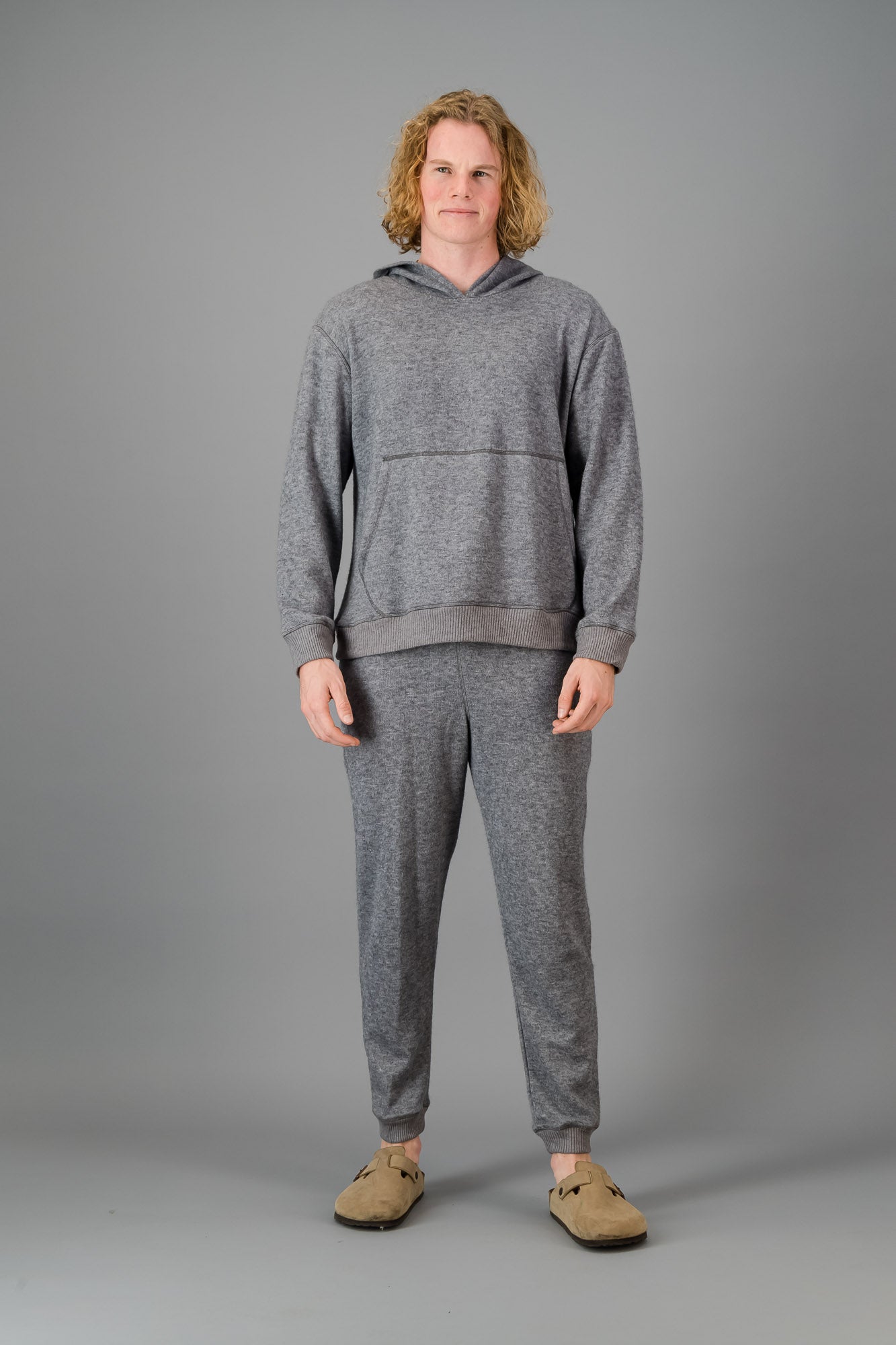 Person with curly blonde hair wearing a comfortable Cashmere-Feel Hoodie (Grey) and matching relaxed fit grey joggers.