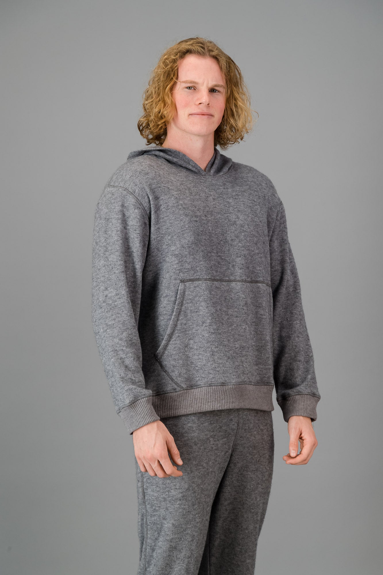 Person with curly blonde hair wearing a comfortable Cashmere-Feel Hoodie (Grey) and matching relaxed fit grey joggers.