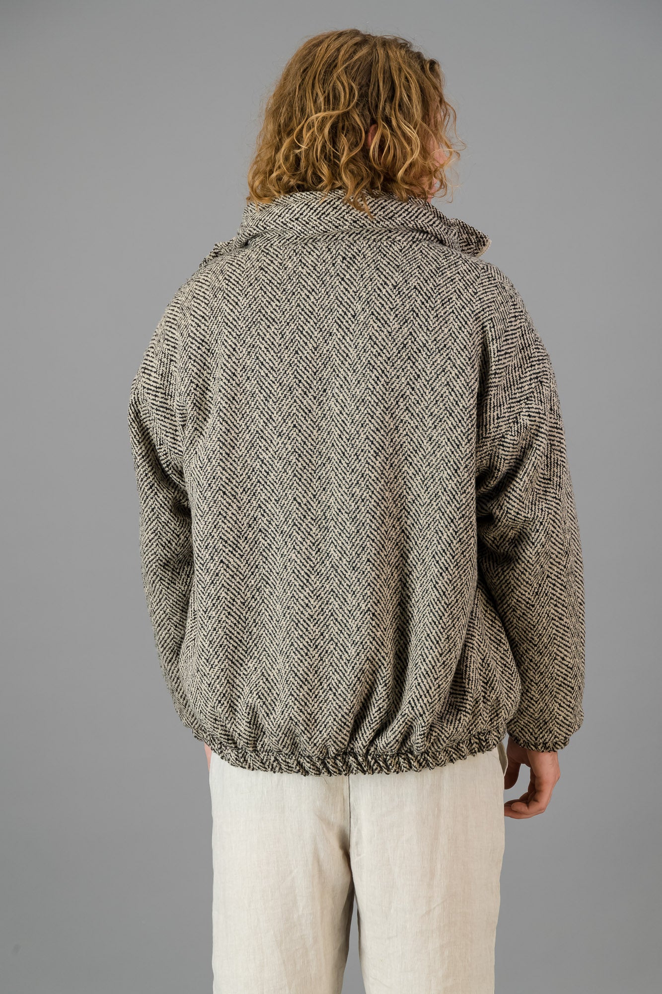 An individual with shoulder-length curly hair is wearing the Herringbone Bomber and light clothing, standing against a gray background.