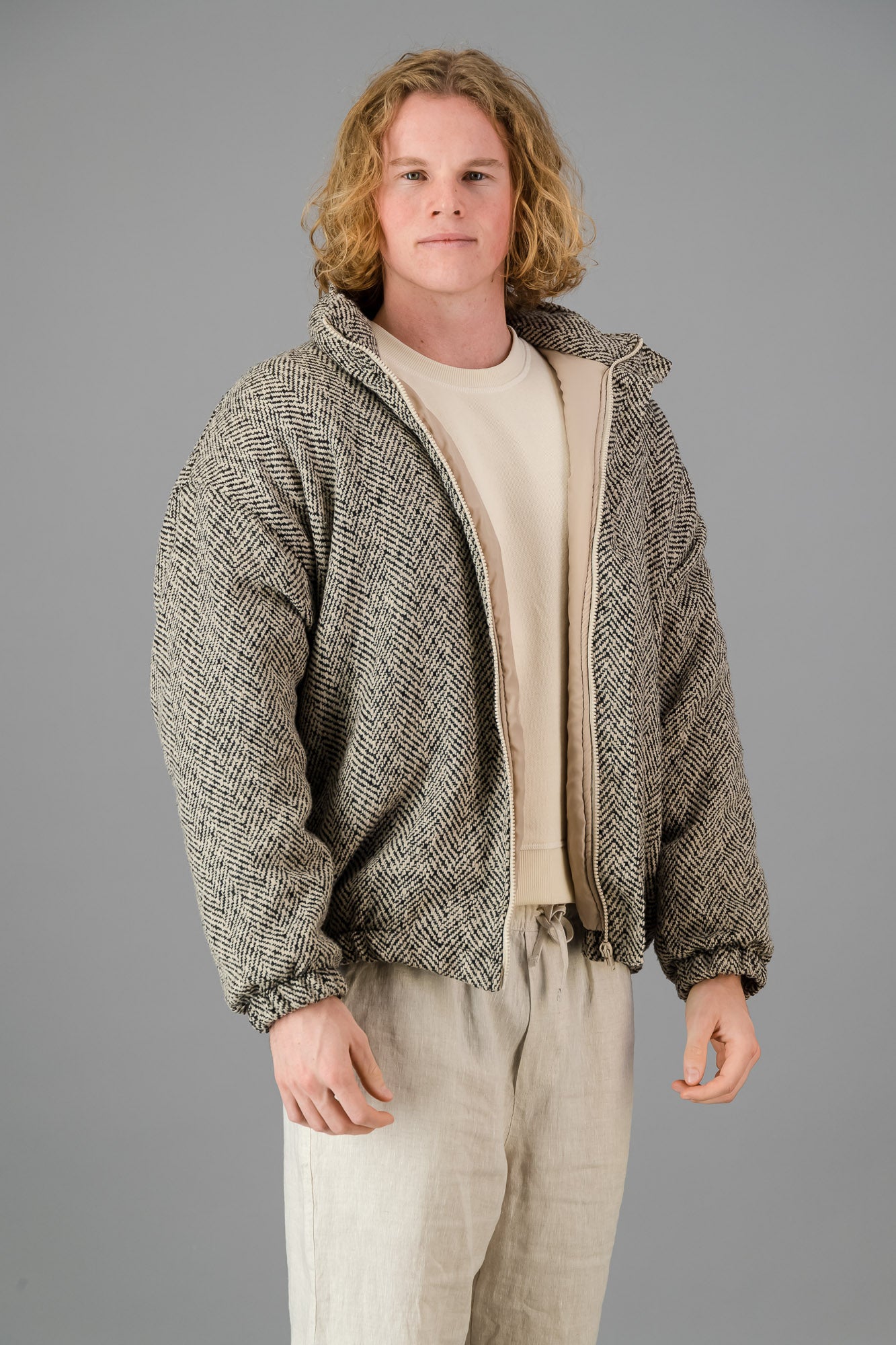 An individual with shoulder-length curly hair is wearing the Herringbone Bomber and light clothing, standing against a gray background.