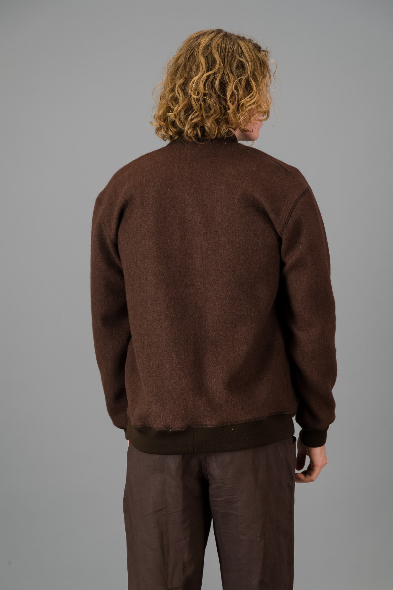 A person with wavy blond hair wearing a versatile layering piece—a chocolate, Knitted Melton Cardigan—over a white shirt and brown pants, standing against a gray background.