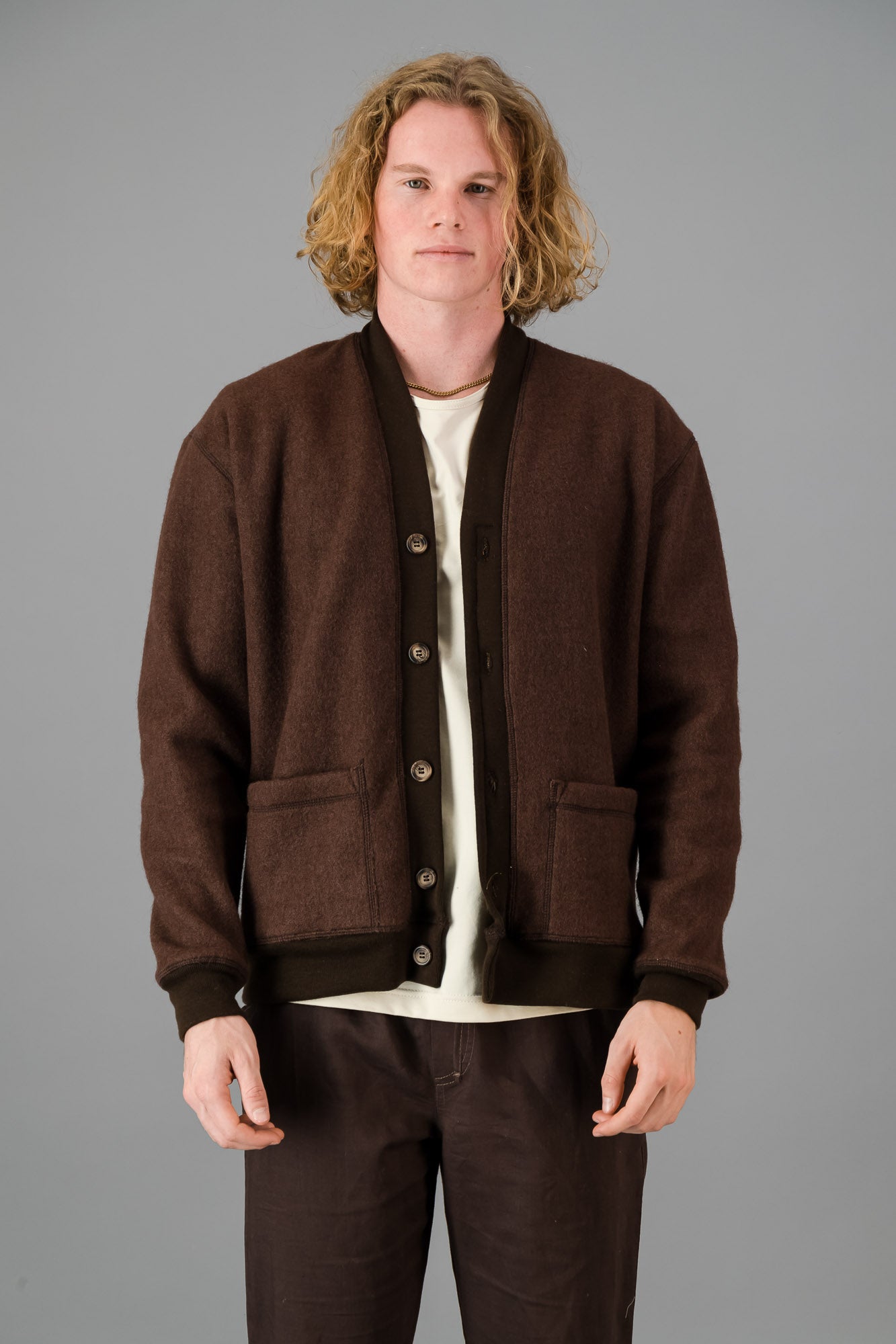 A person with wavy blond hair wearing a versatile layering piece—a chocolate, Knitted Melton Cardigan—over a white shirt and brown pants, standing against a gray background.