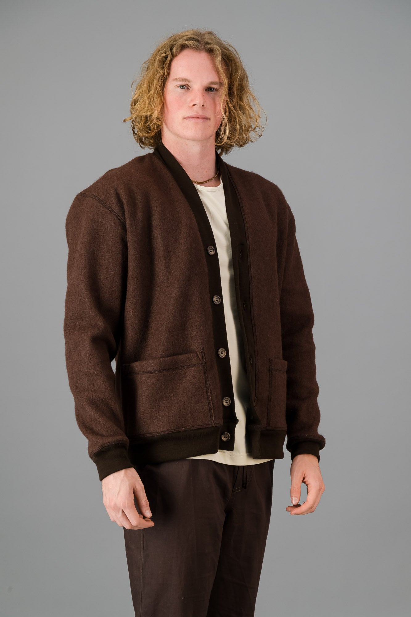 A person with wavy blond hair wearing a versatile layering piece—a chocolate, Knitted Melton Cardigan—over a white shirt and brown pants, standing against a gray background.