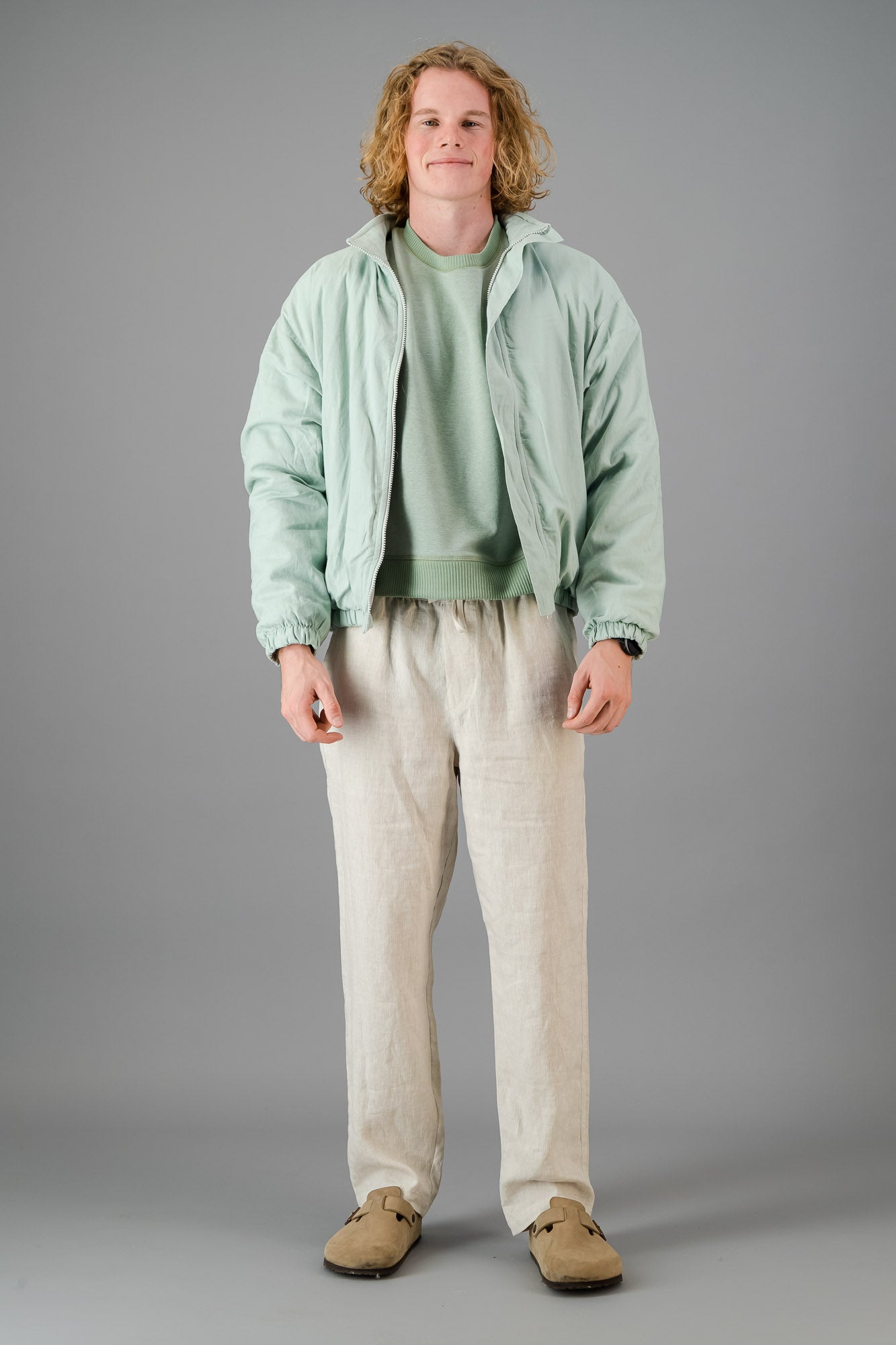 A person with blond, curly hair is wearing a Sage Green Linen Viscose Bomber from the Outerwear Collection over a matching green sweater and light-colored pants, standing against a gray background.