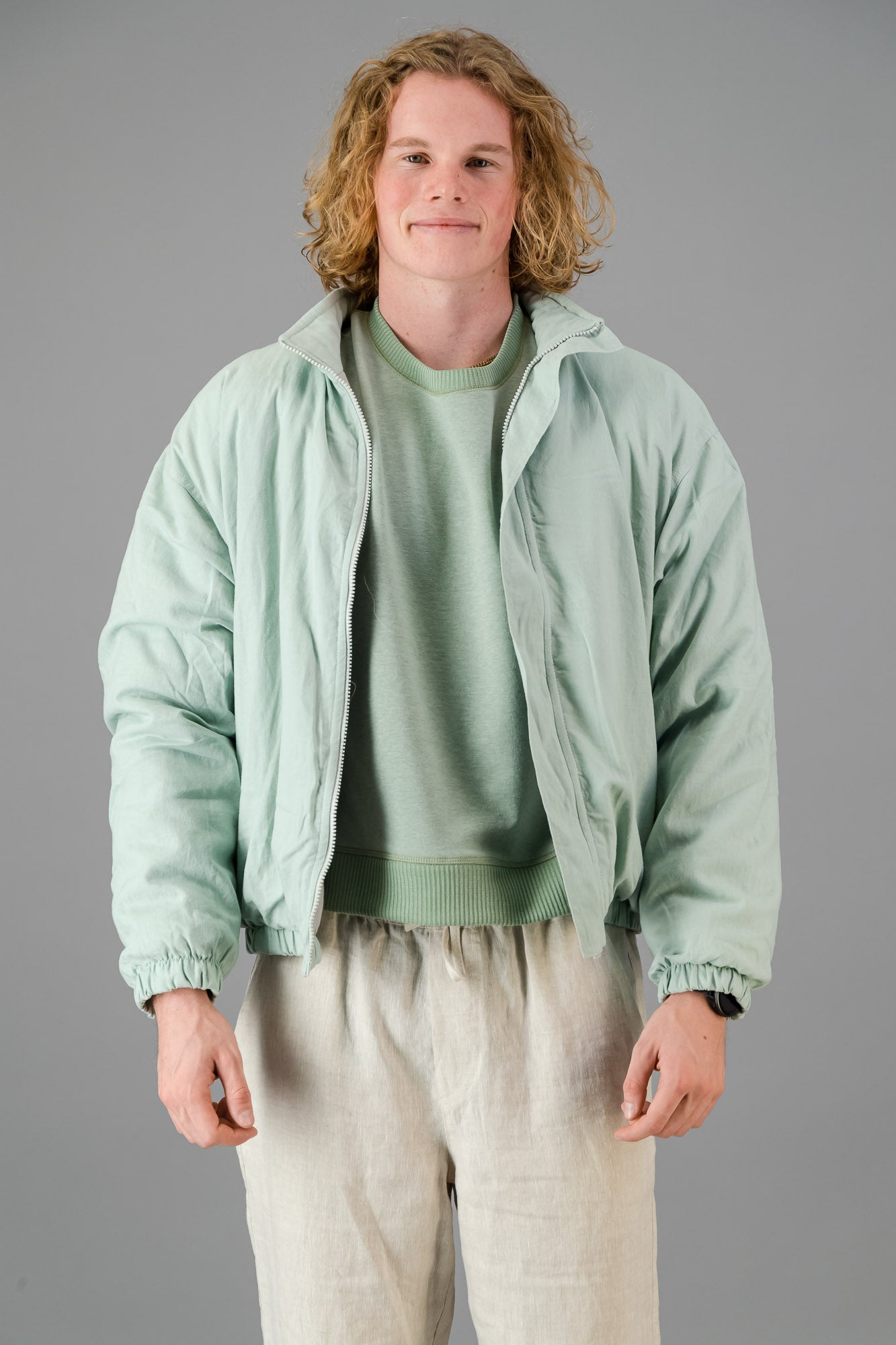 A person with blond, curly hair is wearing a Sage Green Linen Viscose Bomber from the Outerwear Collection over a matching green sweater and light-colored pants, standing against a gray background.