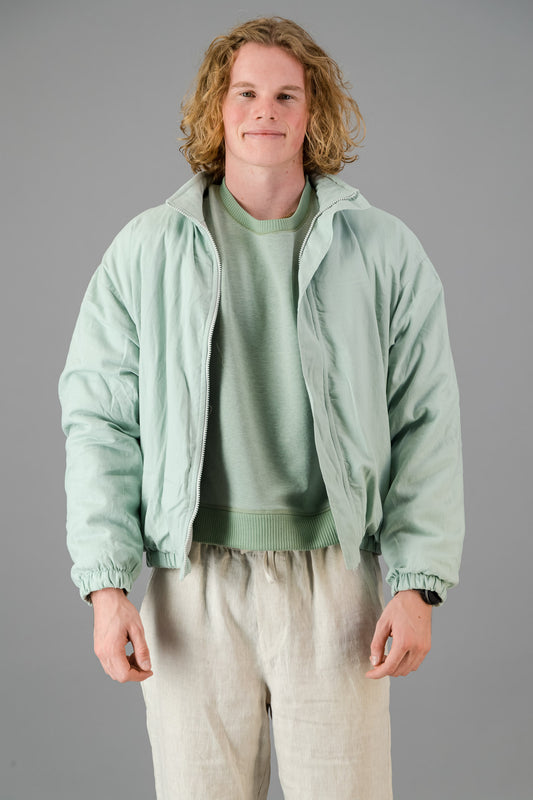 A person with blond, curly hair is wearing a Sage Green Linen Viscose Bomber from the Outerwear Collection over a matching green sweater and light-colored pants, standing against a gray background.