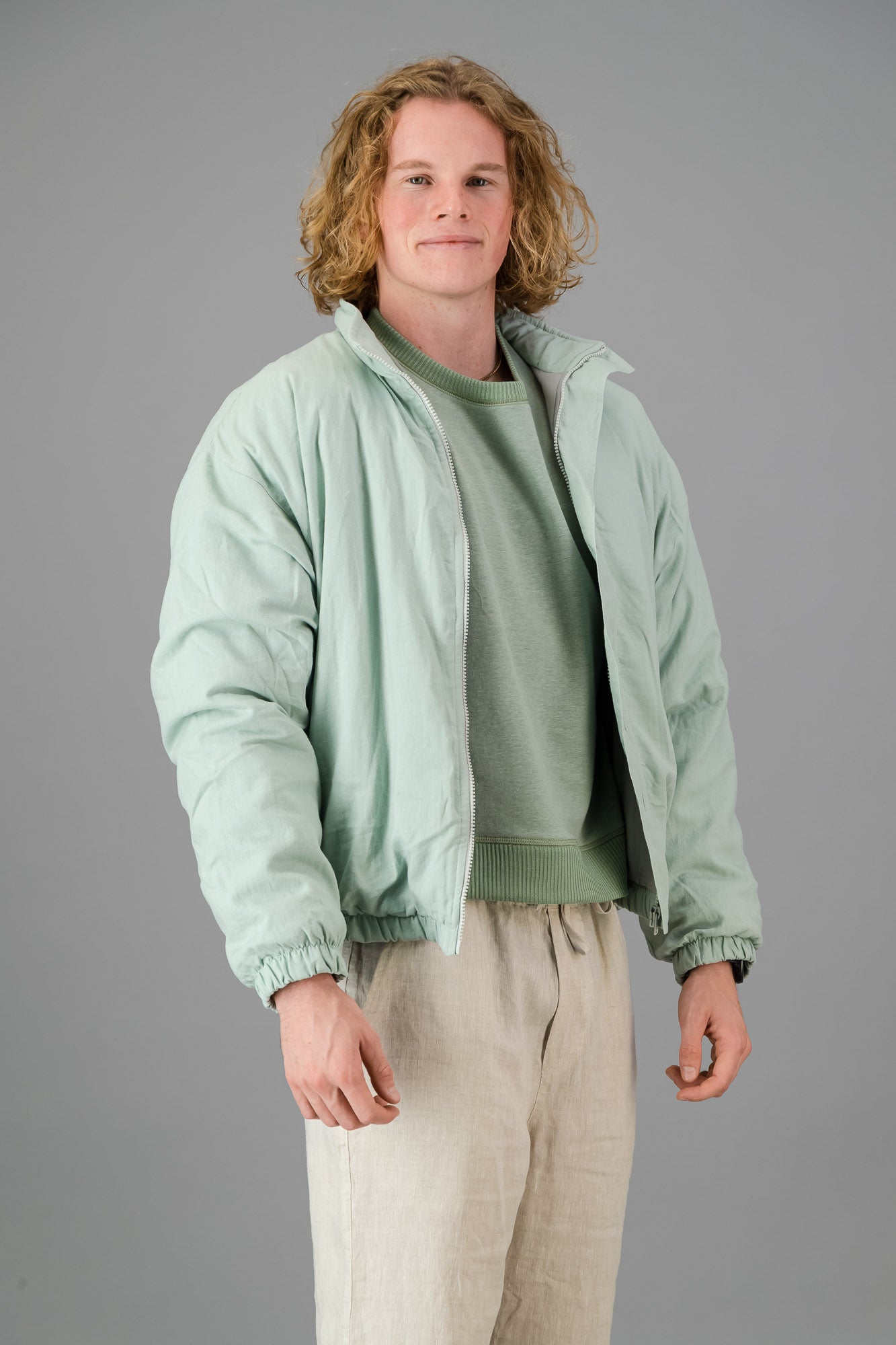 A person with blond, curly hair is wearing a Sage Green Linen Viscose Bomber from the Outerwear Collection over a matching green sweater and light-colored pants, standing against a gray background.