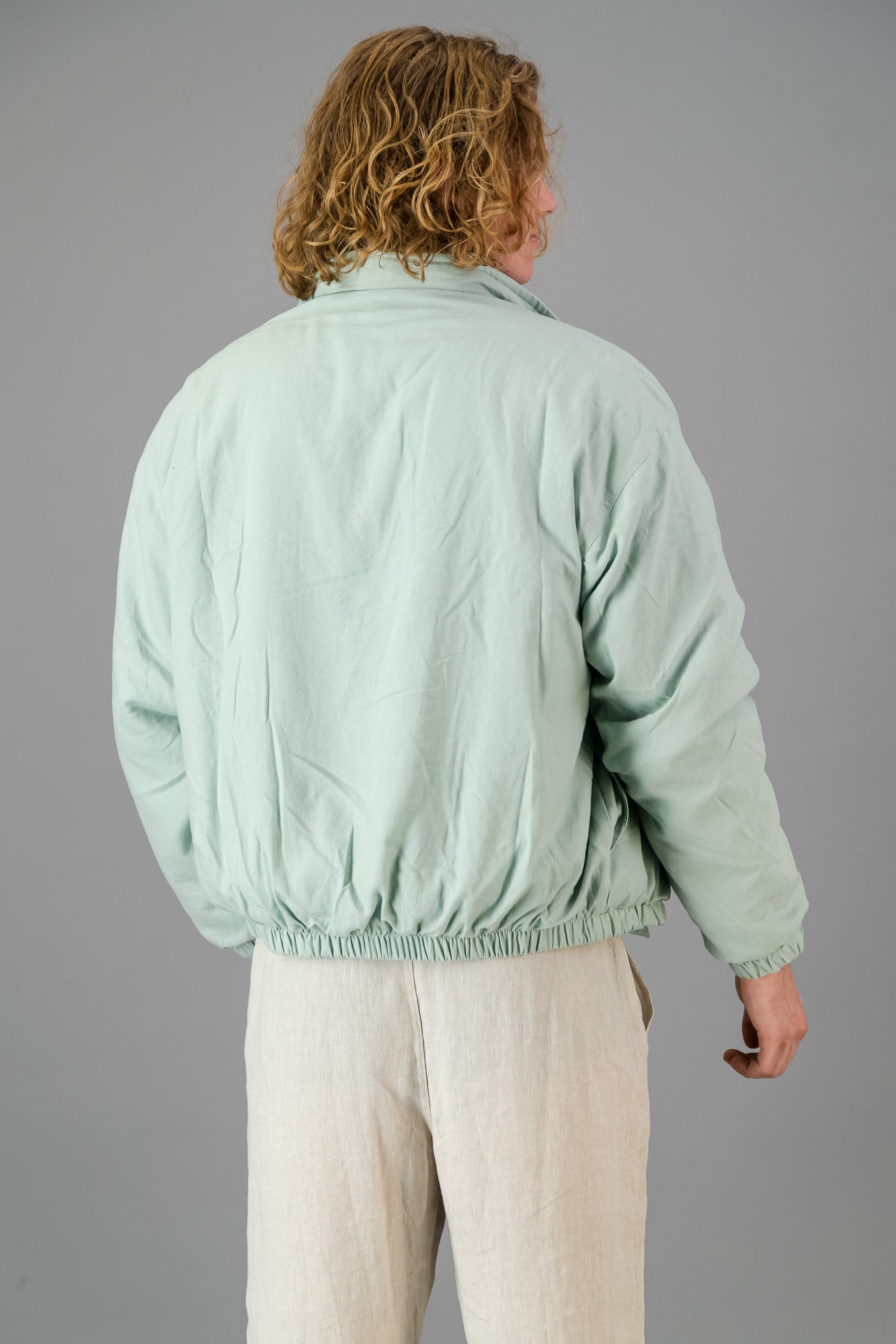 A person with blond, curly hair is wearing a Sage Green Linen Viscose Bomber from the Outerwear Collection over a matching green sweater and light-colored pants, standing against a gray background.