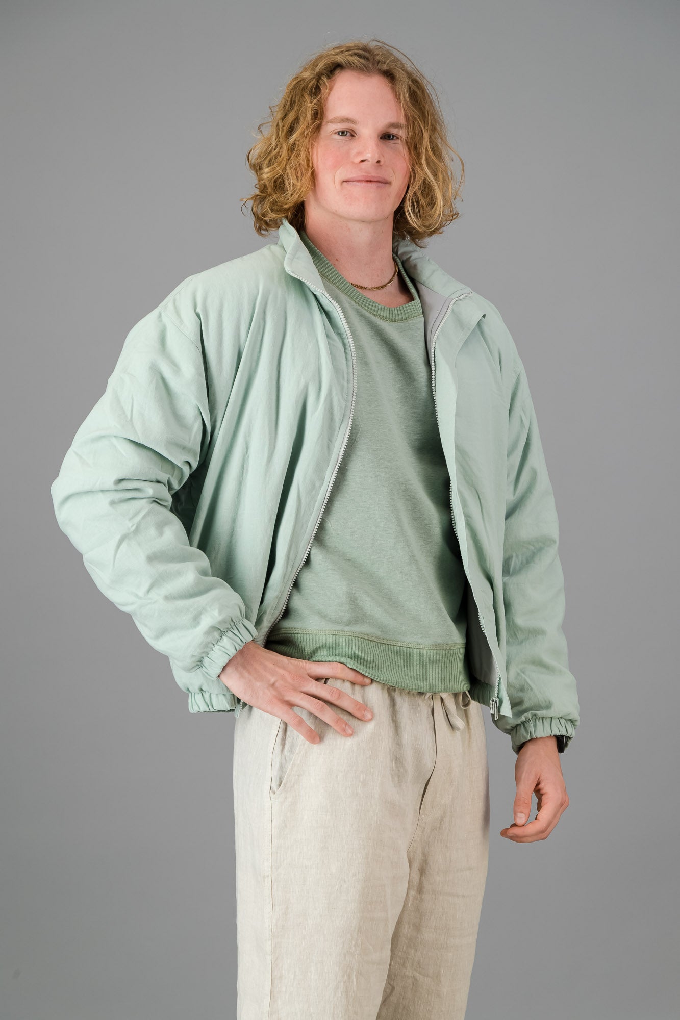 A person with blond, curly hair is wearing a Sage Green Linen Viscose Bomber from the Outerwear Collection over a matching green sweater and light-colored pants, standing against a gray background.