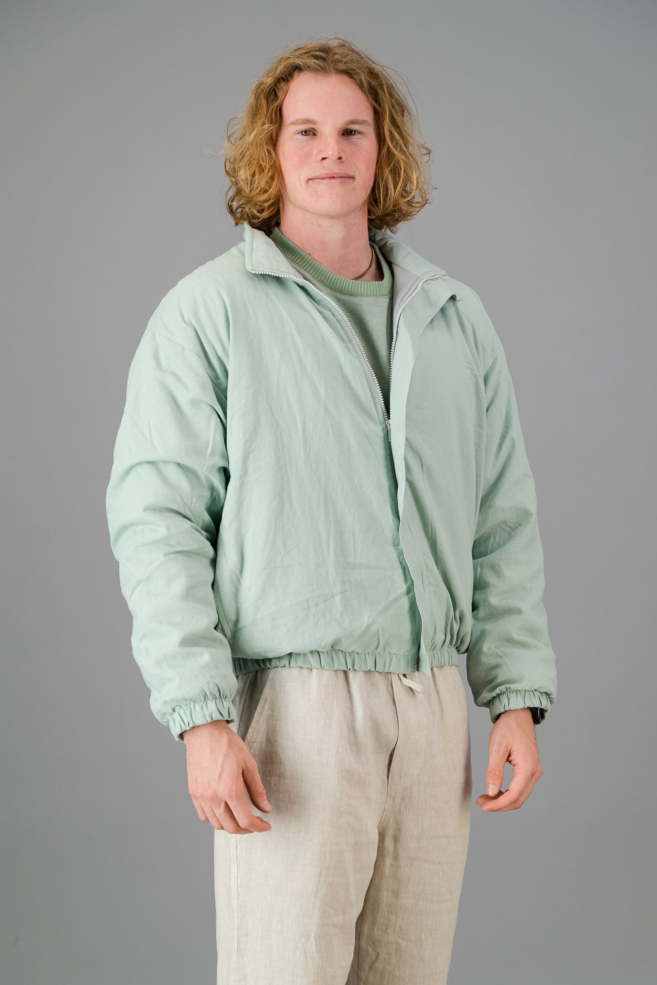 A person with blond, curly hair is wearing a Sage Green Linen Viscose Bomber from the Outerwear Collection over a matching green sweater and light-colored pants, standing against a gray background.