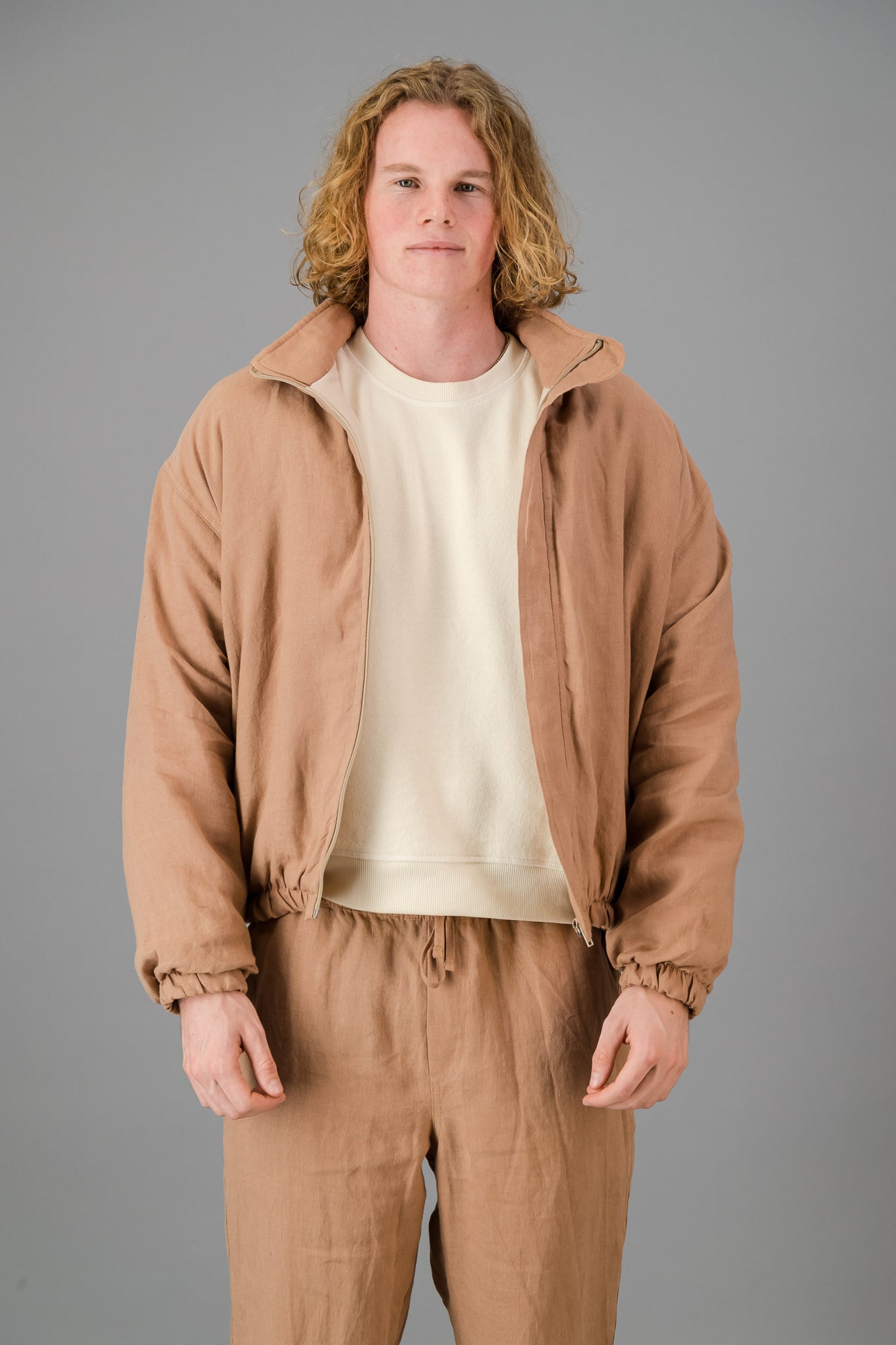 A person with wavy blonde hair wearing the Mocha Linen Bomber over a white shirt and matching beige pants stands against a gray background.