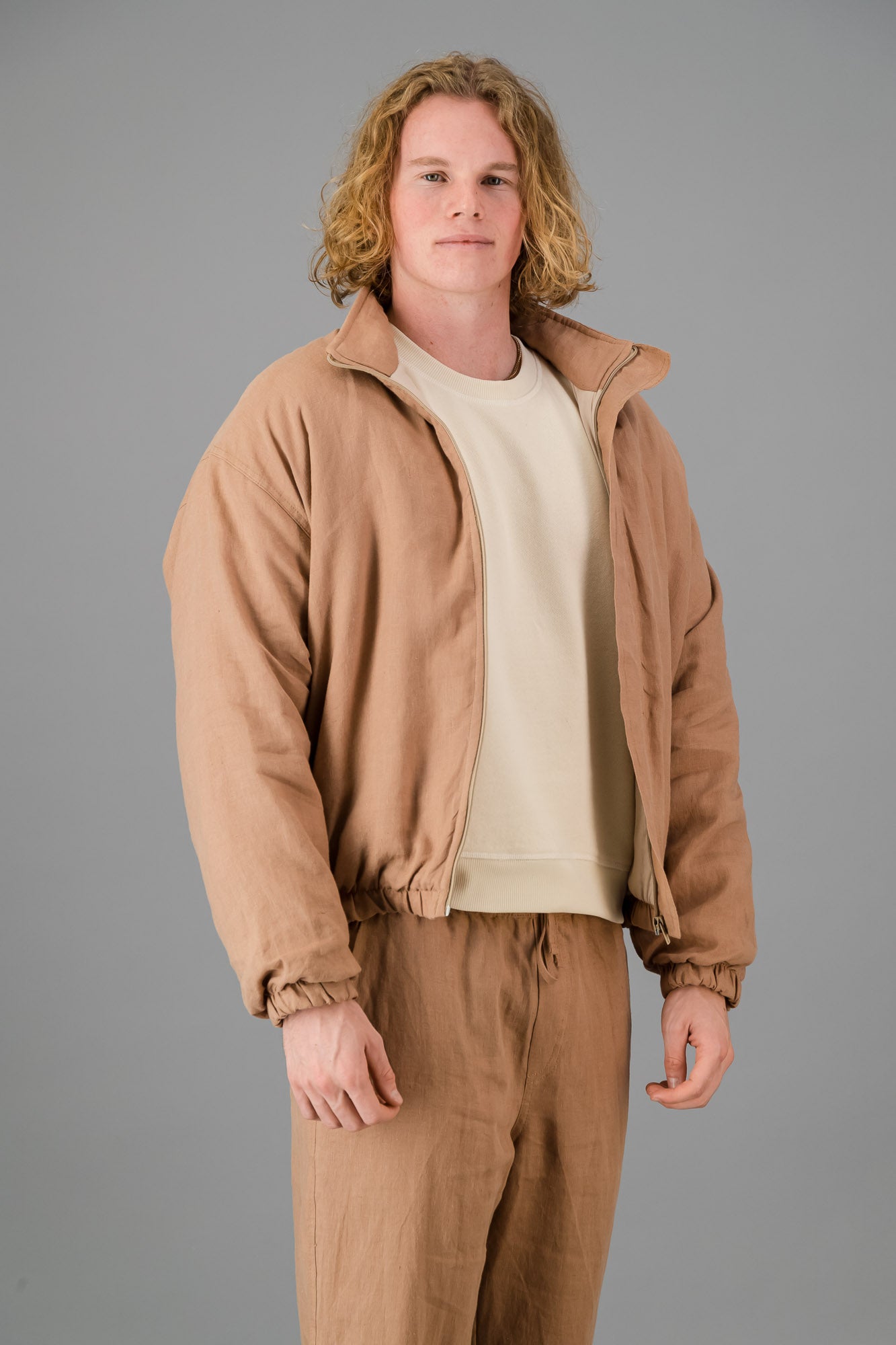 A person with wavy blonde hair wearing the Mocha Linen Bomber over a white shirt and matching beige pants stands against a gray background.