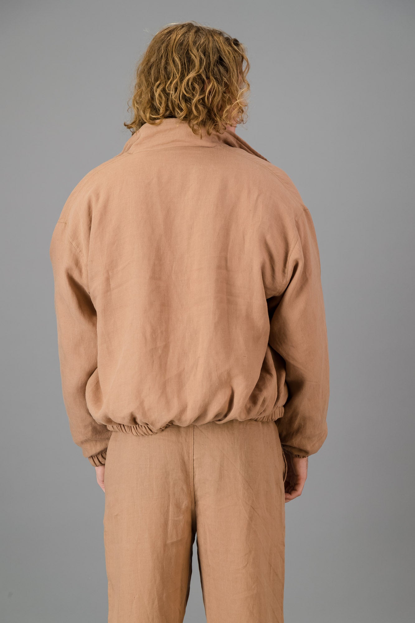 A person with wavy blonde hair wearing the Mocha Linen Bomber over a white shirt and matching beige pants stands against a gray background.