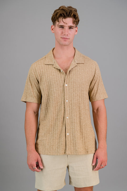 A person with short, curly hair wearing a button-up, short-sleeve Rayon-rich Cable Knit Shirt in brown and oatmeal shorts.