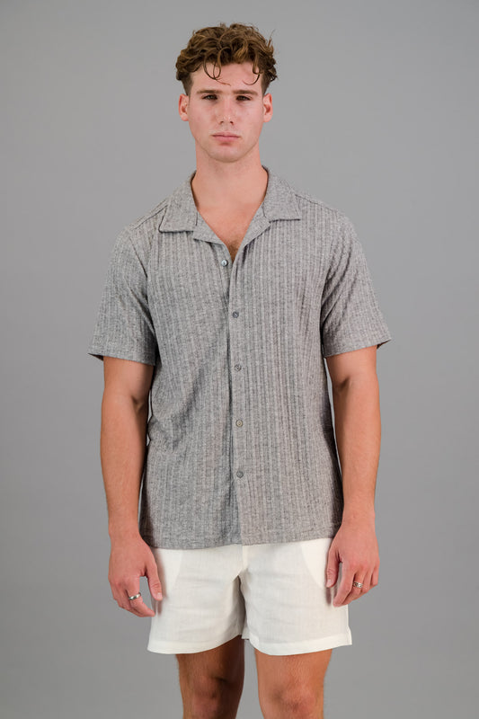 A young man models a Cable Knit Shirt in grey and white shorts against a plain grey background.