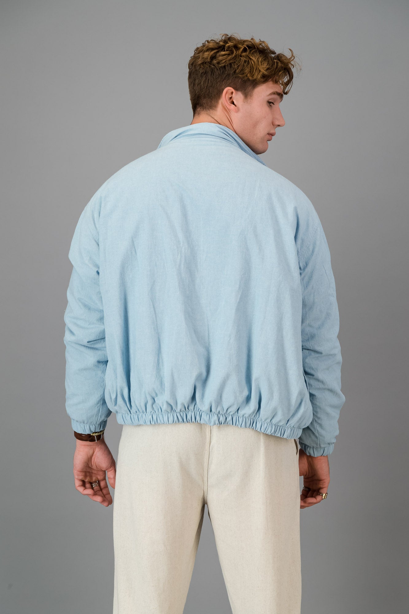 A man with curly hair wears a stylish and cozy Sky Blue Cotton Bomber over a blue shirt with cream pants, standing against a plain grey background.