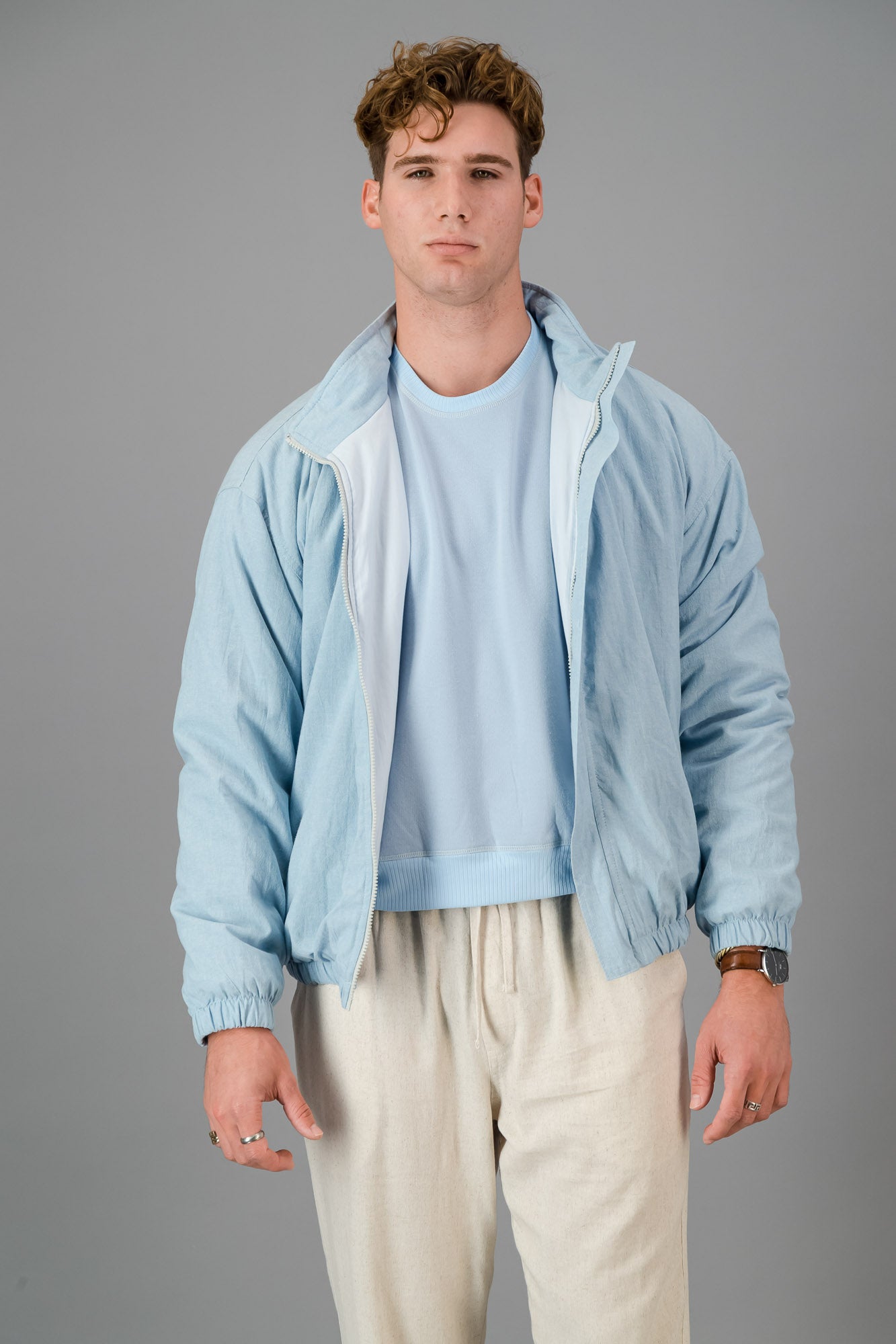 A man with curly hair wears a stylish and cozy Sky Blue Cotton Bomber over a blue shirt with cream pants, standing against a plain grey background.