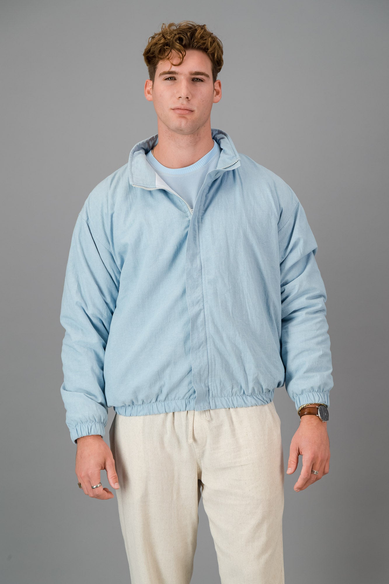 A man with curly hair wears a stylish and cozy Sky Blue Cotton Bomber over a blue shirt with cream pants, standing against a plain grey background.