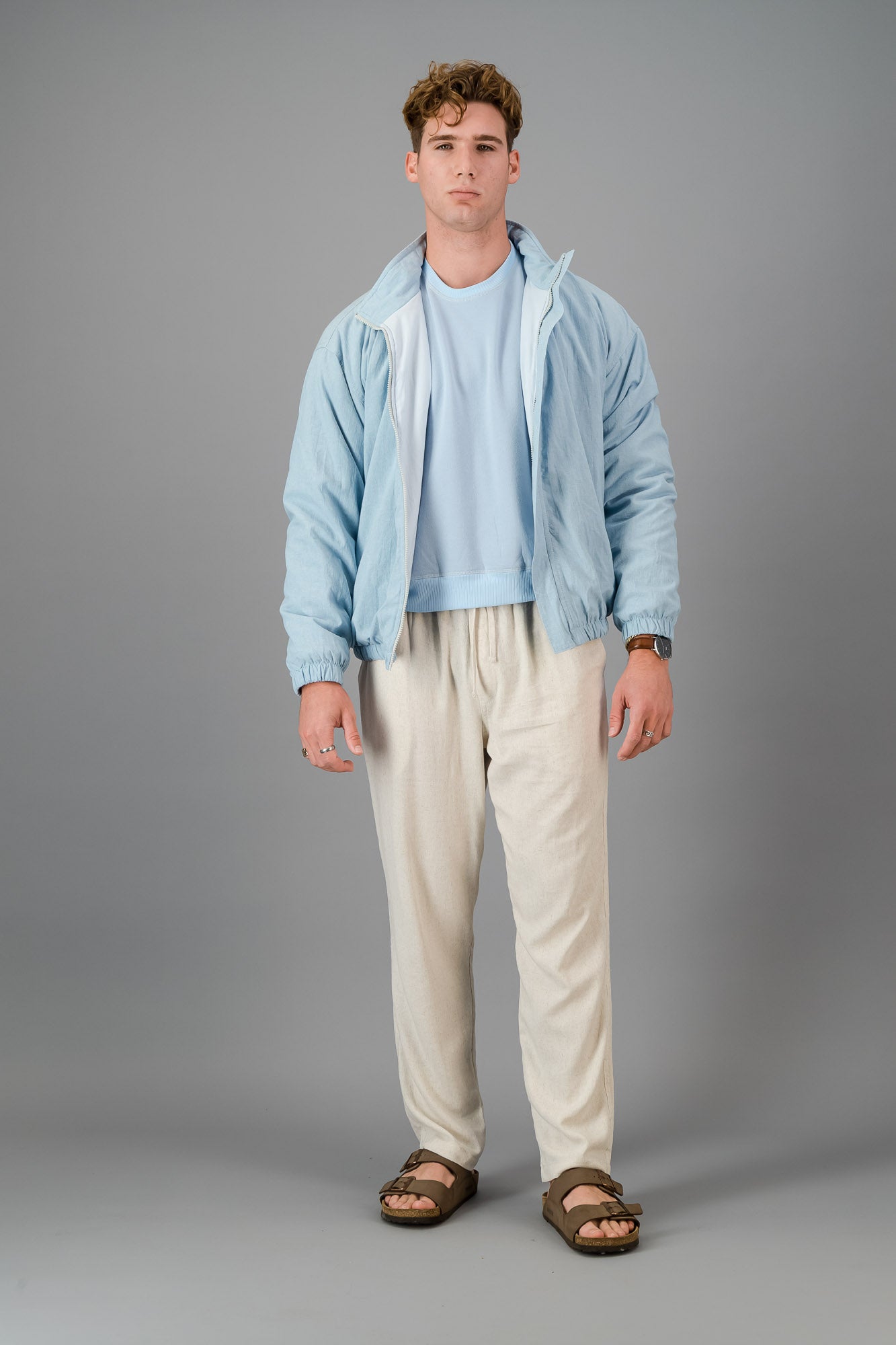 A man with curly hair wears a stylish and cozy Sky Blue Cotton Bomber over a blue shirt with cream pants, standing against a plain grey background.