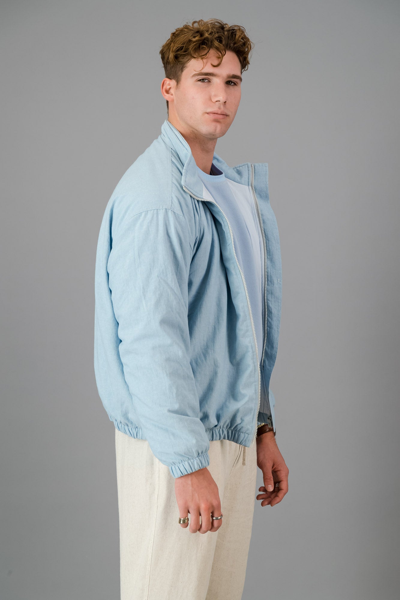 A man with curly hair wears a stylish and cozy Sky Blue Cotton Bomber over a blue shirt with cream pants, standing against a plain grey background.