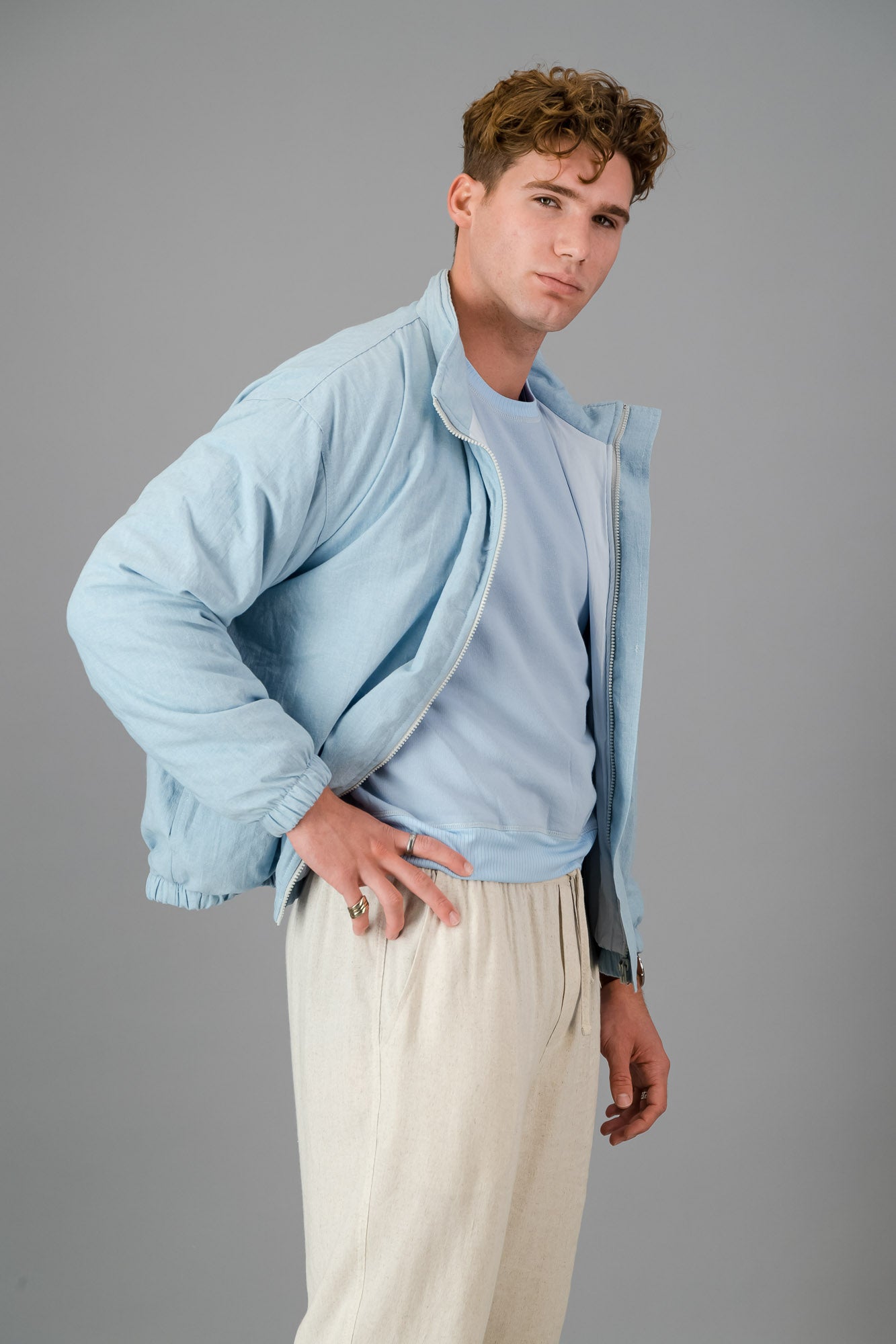 A man with curly hair wears a stylish and cozy Sky Blue Cotton Bomber over a blue shirt with cream pants, standing against a plain grey background.