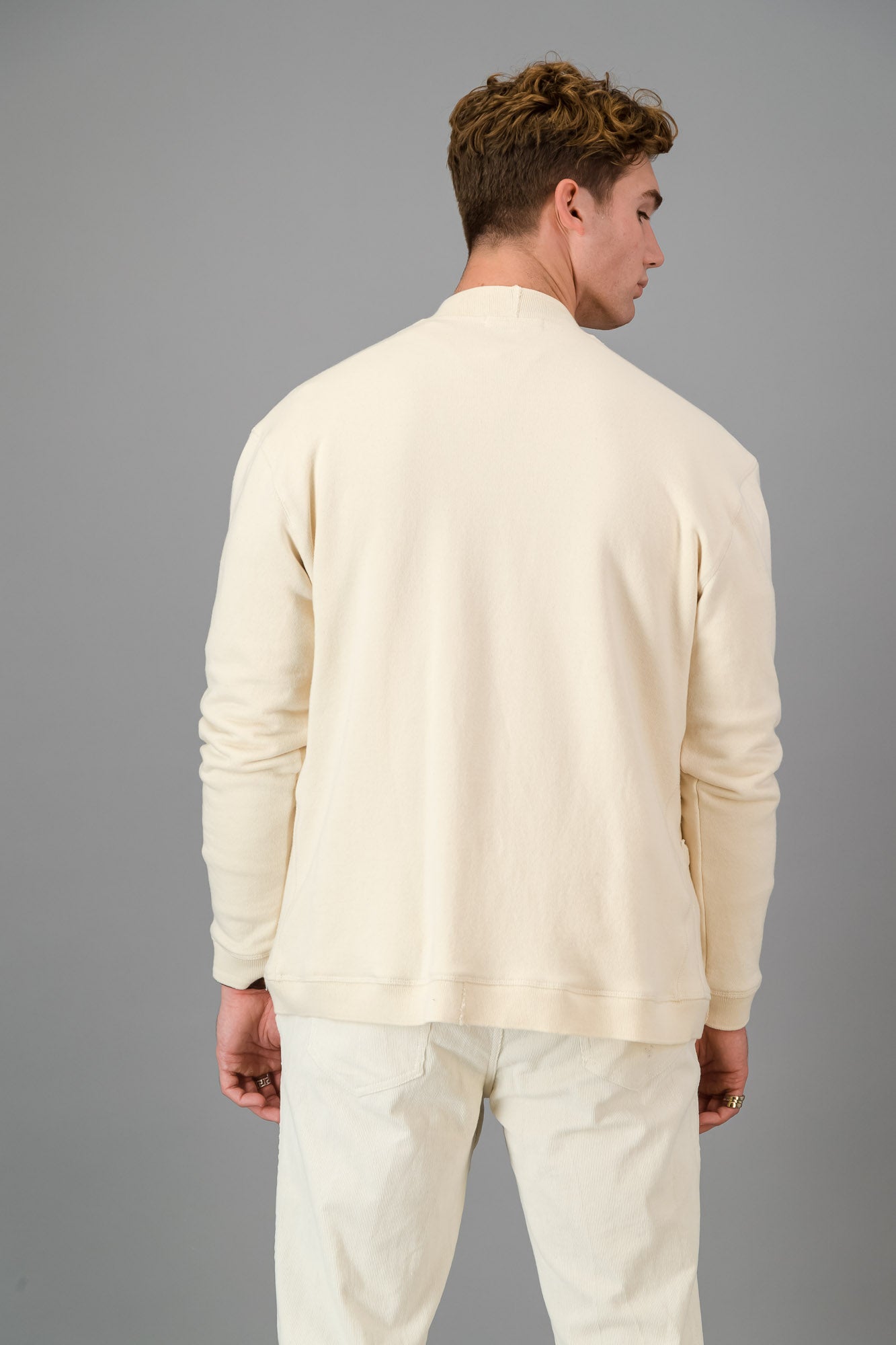 Man in a cream-colored outfit, showcasing the Vanilla Cotton Cardigan—a versatile layering piece known for its luxurious softness—comprising a button-up cardigan, T-shirt, and pants, standing against a gray background.