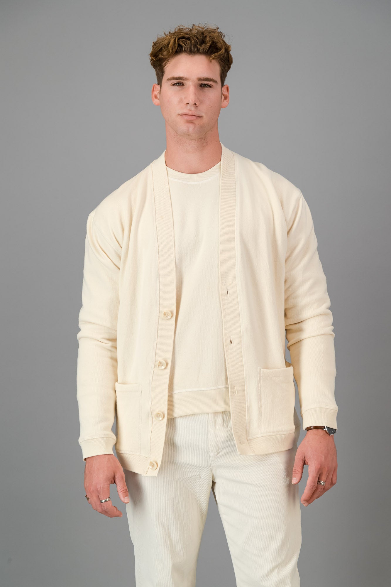 Man in a cream-colored outfit, showcasing the Vanilla Cotton Cardigan—a versatile layering piece known for its luxurious softness—comprising a button-up cardigan, T-shirt, and pants, standing against a gray background.