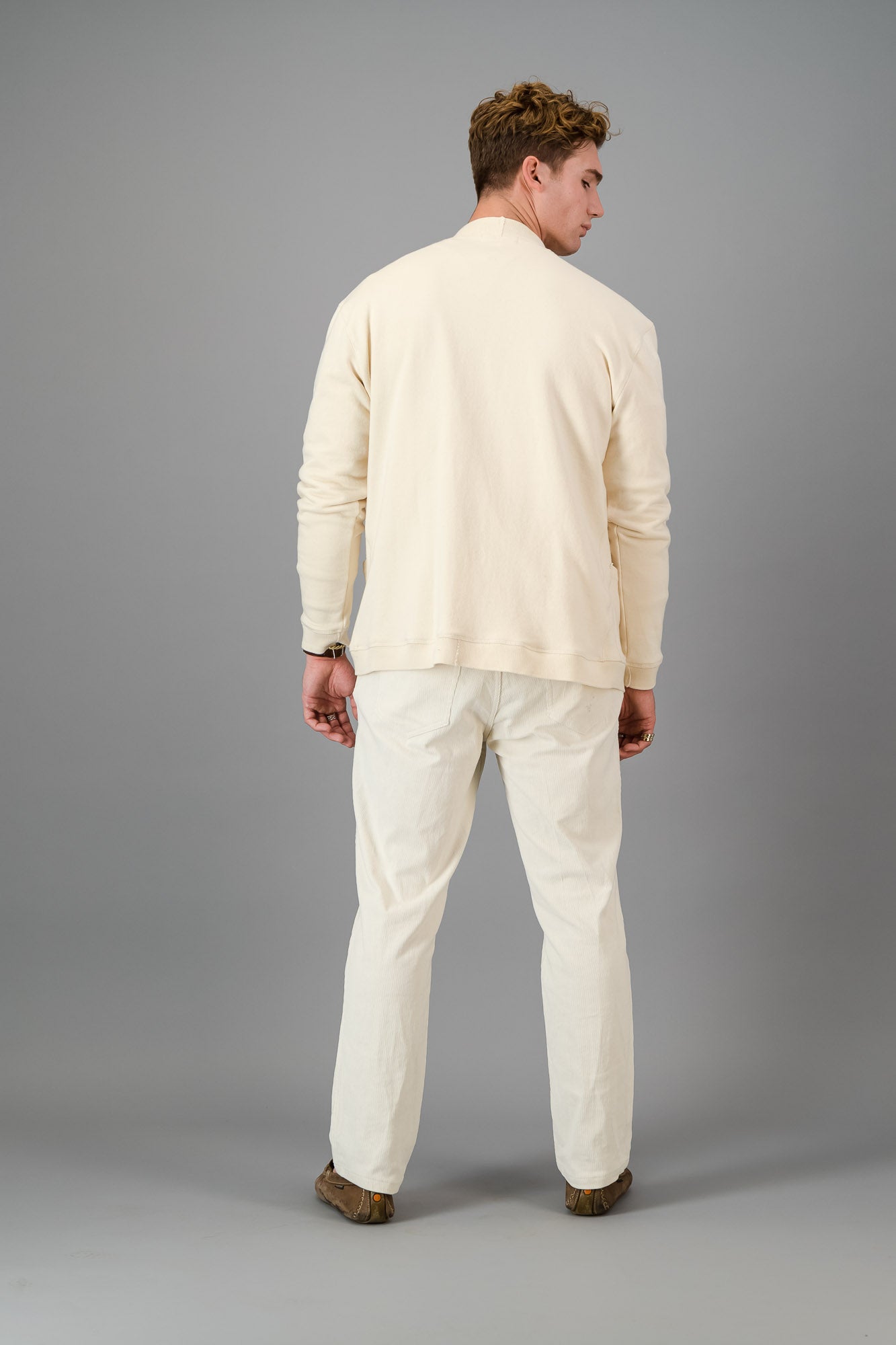 Man in a cream-colored outfit, showcasing the Vanilla Cotton Cardigan—a versatile layering piece known for its luxurious softness—comprising a button-up cardigan, T-shirt, and pants, standing against a gray background.