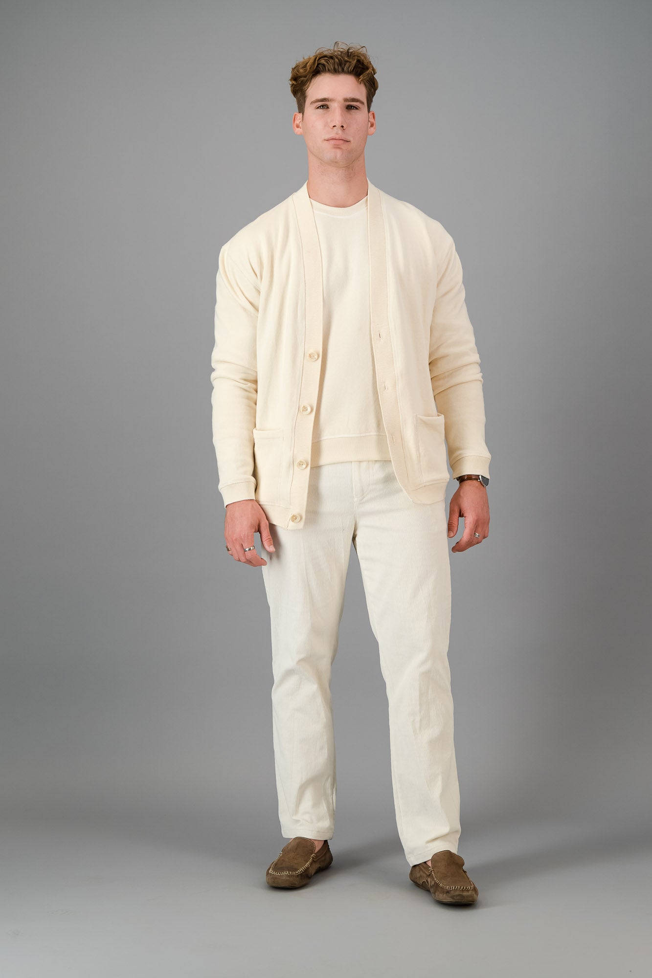 Man in a cream-colored outfit, showcasing the Vanilla Cotton Cardigan—a versatile layering piece known for its luxurious softness—comprising a button-up cardigan, T-shirt, and pants, standing against a gray background.