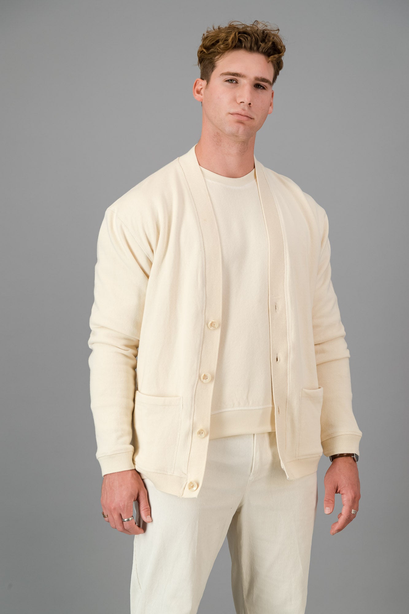 Man in a cream-colored outfit, showcasing the Vanilla Cotton Cardigan—a versatile layering piece known for its luxurious softness—comprising a button-up cardigan, T-shirt, and pants, standing against a gray background.